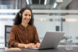 © Liubomir - Woman wearing headphones smiling while typing on a laptop, connecting remotely for a business meeting or online work in a modern corporate office environment