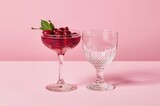 A cocktail glass filled with fresh cherries next to an empty glass stand on a table against a soft pink backdrop with ample space for text and design