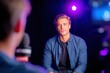 © Natalia - Male actor seated for an interview in a studio setting, with colorful lighting and camera equipment visible, capturing the essence of a professional media interaction