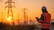 © EarthWalker - Electrical engineer technician in hard hat with a tablet computer device monitoring high voltage transmission tower power grid lines at sunset
