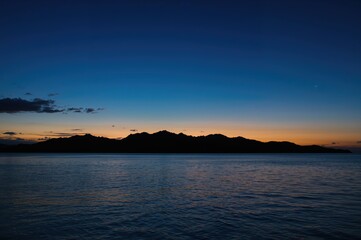 Wall Mural - Tranquil twilight on a tropical isle with mountain shadows set against a vibrant blue and orange sky, mirrored by calm ocean waters.