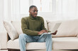 © SHOTPRIME STUDIO - Young Black man sitting comfortably on a couch, using a laptop for work or study in a cozy indoor atmosphere