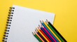 © Ilham - Top-down composition showcasing colored pencils resting near a blank, grid-patterned notebook on a vivid yellow background, flat lay, bright light, studio.