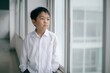 © id512 - Thoughtful young asian schoolboy standing by window in white shirt indoors. Elementary male student looking outside in hallway of modern school building
