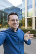 Man recording himself with a smartphone, making a video call, or streaming live in an urban environment with modern office buildings, actively gesturing and smiling