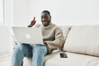 © SHOTPRIME STUDIO - Happy young Black man working on a laptop at home, wearing a cozy sweater and showing a thumbs up while sitting on a light colored sofa