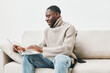 © SHOTPRIME STUDIO - Young African man wearing a cozy sweater, joyfully working on a laptop on a sofa in a minimalist living room, emphasizing productivity and comfort