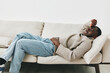 © SHOTPRIME STUDIO - Young Black man in casual attire relaxed on a sofa, expressing emotions of stress and discomfort while holding his head
