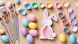 © AliRza - An overhead shot of a wooden table covered with Easter art supplies, including painted eggs, paintbrushes, paint pots, and a cardboard bunny cutout. The image highlights a fun holiday craft activity.