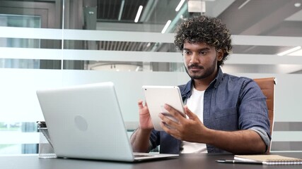 Focused businessman is using digital tablet while sitting at desk at workplace in business office. Handsome manager is engaged in a task while working on a computer, typing message or chatting online