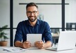 © Alif - Smiling businessman in blue shirt and glasses using a tablet at his desk with laptop and papers for modern office work.