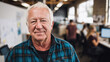 © Som.Ying - Senior man wearing plaid shirt smiling in modern office environment with colleagues working at computers in the background and natural light coming through large windows.