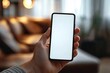 © 宜然 左 - Hand holding a smartphone with a blank white screen in a cozy indoor environment with warm lighting and blurred background