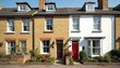 Terraced houses with yellow brick, white walls stand under bright sunny day. Front gardens bloom with colorful flowers, green plants. Traditional UK architecture with dormer windows, distinct doors