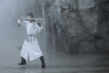 Man in a white robe practicing martial arts with a sword outdoors in a misty environment.