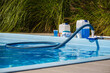 © StockMediaSeller - Swimming pool vacuum hose and brush cleaning water, with maintenance products including chemical canisters, chlorine tablets, dispenser, measuring cup and test kit placed on poolside