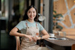 © PaeGAG - Smiling Asian woman barista wearing an apron, sitting at a cafe table with a cup of coffee and smartphone
