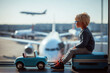 © Giovanna - Boy with blond hair and glasses sits on a blue suitcase at the airport window, with a turquoise toy car in front, watching planes outside. Perfect for travel, tourism, vacation, childhood lifestyle