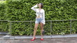 © iFocus - Asian female model stands outdoors in front of a green hedge, leaning against a railing in red heels, denim shorts, and an unbuttoned white shirt
