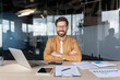 © Liubomir - Confident young businessman seated at desk with arms crossed, smiling at camera while laptop, documents and tablet suggest productive modern office work and leadership