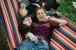 © qunica.com - Two friends enjoy a peaceful moment outdoors, reclining on a striped hammock in a lush green park, sharing laughter and experiencing joy under the warm sunlight.
