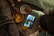 © DragonImages - Black man and Black child sitting on floor watching movie on tablet, eating popcorn and chips, drinking juice, child wearing bracelets, tablet displaying outdoor scene with cityscape