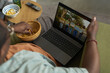 © DragonImages - Black man sitting on sofa eating chips while watching laptop screen showing group of diverse young adults holding signs outdoors, remote and snacks on table nearby
