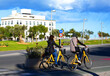 Two cyclists ride yellow bikes past modern building on sunny day in coastal city. People cycle on yellow bikes alongside park area. Scene captures city life and outdoor activity in Valencia, Spain.