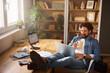 © Stockphotodirectors - A man with curly hair drinks coffee while sitting comfortably in a chair, using a laptop in a bright home office filled with greenery and shelves.