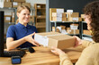 © pressmaster - Caucasian young adult woman smiling while handing package to customer across counter in shipping or postal service office, shelves with parcels visible in background, barcode scanner on desk
