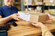 © pressmaster - Caucasian woman handing cardboard package to another woman across wooden counter in shipping or delivery service office, both women interacting with parcel during transaction