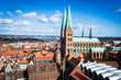 Vista aérea de la ciudad de Lübeck con sus característicos tejados rojos. En el centro, destaca la imponente Iglesia de Santa María con sus dos altas agujas verdes bajo un cielo azul despejado.