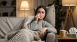 © ARI - A woman sits on a couch, wrapped in a blanket, with a tissue in her hand, appearing sad and unwell in a dimly lit living room.