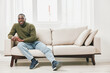 © SHOTPRIME STUDIO - Smiling man sitting on a sofa in a stylish living room, wearing a green sweater and jeans, conveying warmth and comfort, ideal for lifestyle themes