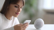© Allie - Woman using smartphone near smart speaker on table