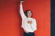 © BullRun - Young happy woman enjoying music in headphones, raising phone, smiling with joy against red wall, symbolizing connection, entertainment, and emotional expression.