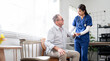 © NAMPIX - A friendly female nurse in blue scrubs stands beside an elderly male patient, offering him comfort and support. The clinic or home setting suggests a moment of compassionate healthcare,