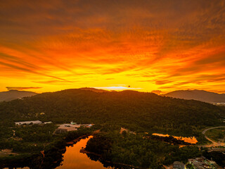 Wall Mural - A breathtaking aerial view of a vivid orange sunset casting dramatic light over forested hills and a reflective river, with urban elements nestled at the landscape’s edge beneath a glowing sky.
