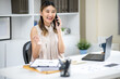 © bongkarn - Pretty woman office worker raising hand holding pen while talking on phone at white table in office.