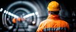 © Alohanongza - A construction worker in a hard hat and orange jacket stands in a brightly lit underground tunnel, focused on the work ahead