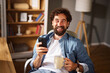 © Stockphotodirectors - A man with curly hair and a beard sits in a chair, holding a mug of coffee in one hand and a smartphone in the other, smiling in a comfortable home office environment.