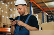 © standret - Uses smartphone, holding it in hands. Man in uniform is working in the storage with packages for shipment