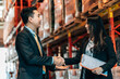 © BigBlues - A businessman and businesswoman in suits are shaking hands in a warehouse aisle. The woman is smiling and holding a clipboard.