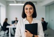 © shahida  - Confident young professional woman smiling holding tablet in modern office with colleagues in background