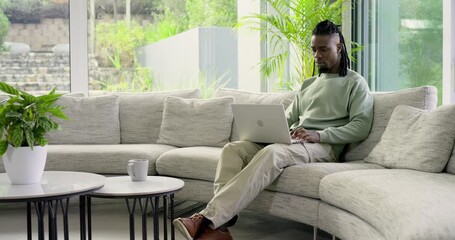 African American man pausing typing on sofa viewing garden resuming laptop work at home, copy space