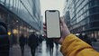 © Hasriana - A hand in a yellow jacket holds a smartphone with a blank screen, outdoors in a blurred city street on a cloudy day. People are walking in the background