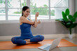 © Pang - A young woman practicing yoga indoors on an exercise mat, using a laptop for online training, symbolizing health, strength, and wellbeing.
