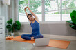 © Pang - A young woman practicing yoga indoors on an exercise mat, using a laptop for online training, symbolizing health, strength, and wellbeing.
