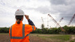 © Happy Photo - A worker in an orange safety vest and helmet watches cranes at a construction site near a rural area. The sky is cloudy, hinting at possible rain