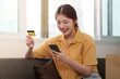 © Natee Meepian - Online Shopping. Young woman using smartphone and credit card for online purchases at home.